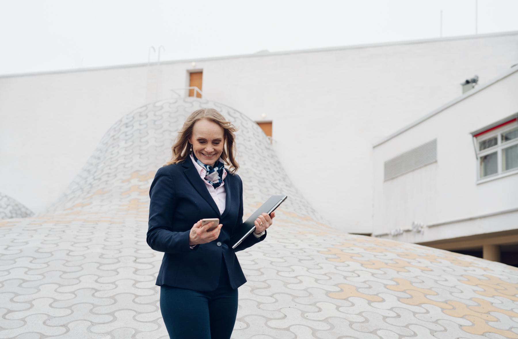 Nainen hymyilee ja selaa puhelinta kannettava tietokone kainalossaan Helsingin keskustassa.